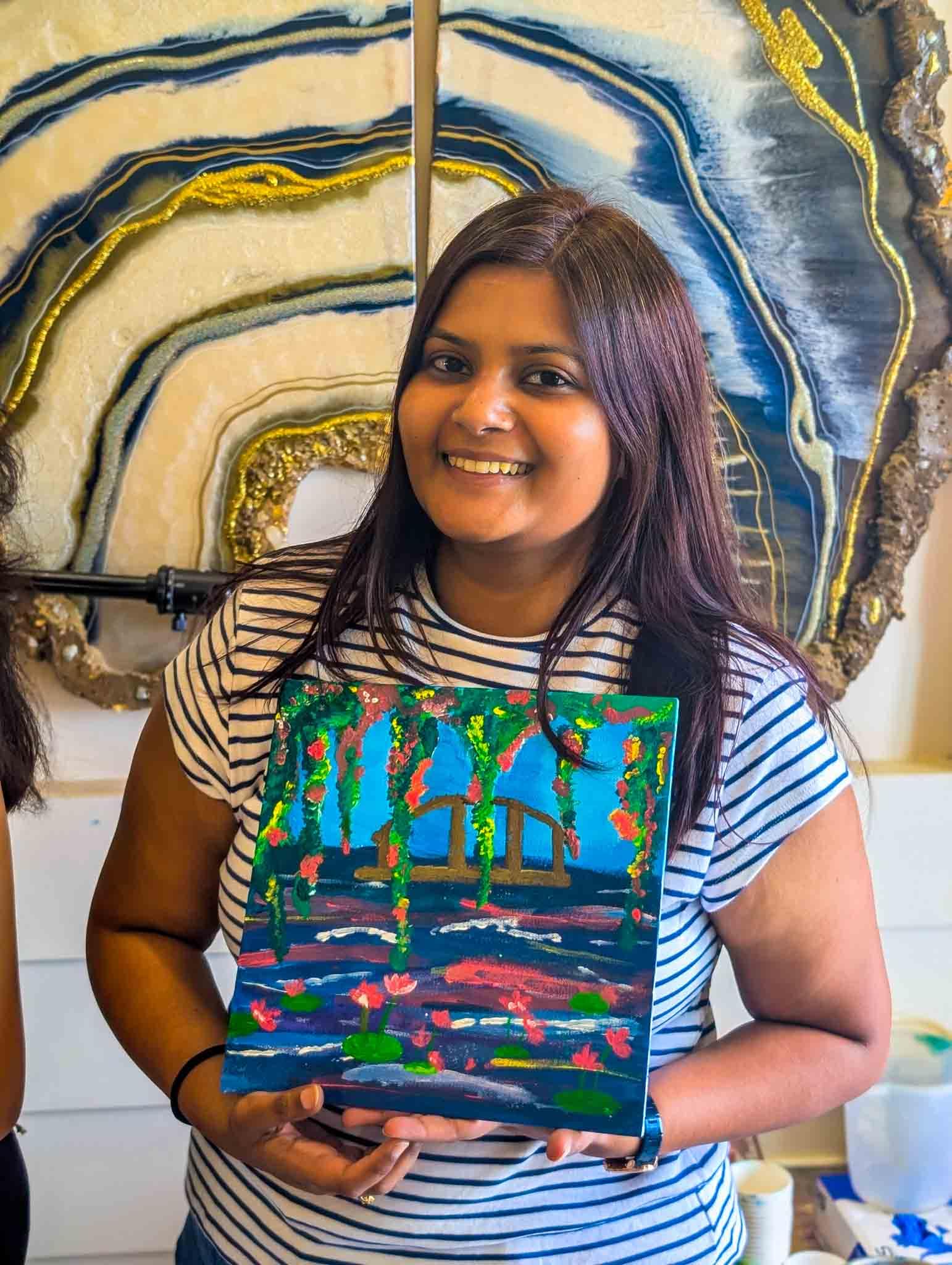 Happy participant displaying her painting of a peaceful blue water scene with a bridge and hanging flowers in a Sip & Paint event.
